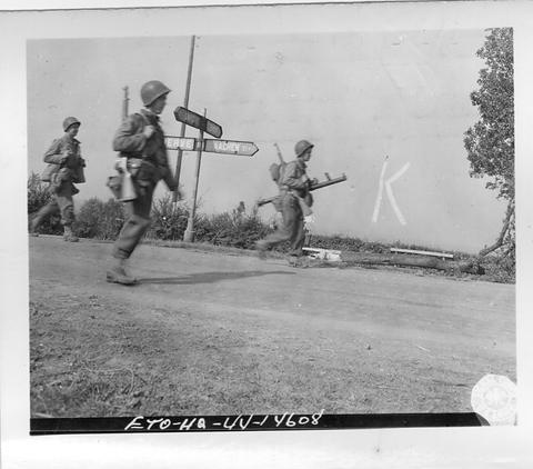 Period photograph: three American soldiers walking along a road past a signpost, one carrying a bazooka, a letter K painted on the wall behind