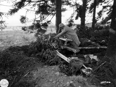 Period photograph: a damaged anti-tank gun under fir branches, a soldier bending over it, equipment on the ground