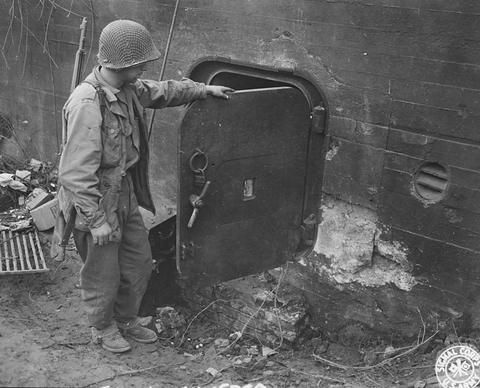 Period photograph: a soldier in a helmet pointing at a steel door set in a concrete wall, rubble at its foot