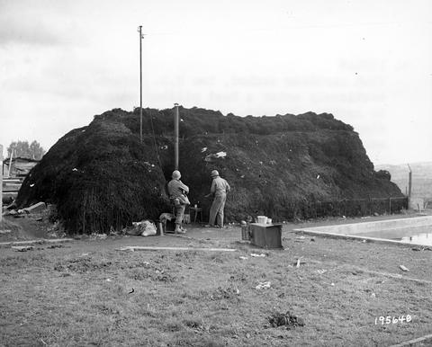 Period photograph: a large mound covered with earth and turf, two soldiers at its side, factory buildings behind