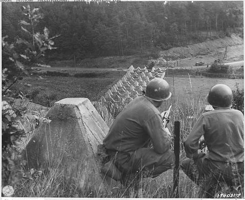 Period photograph: two helmeted soldiers seen from behind, crouching above a row of concrete obstacles in a valley
