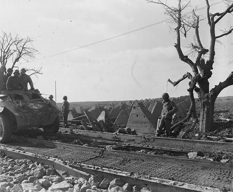 Period photograph: an armoured car on a track beside lines of concrete tank obstacles, a soldier standing nearby