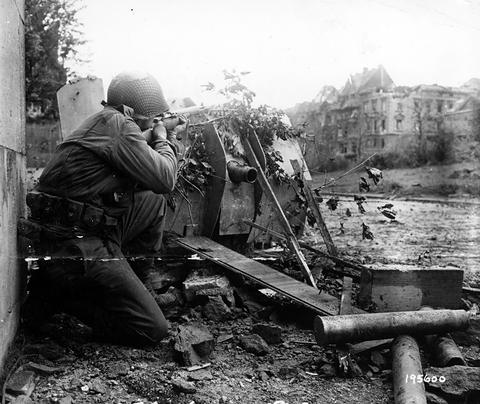 Period photograph: a soldier crouching with a rifle behind the shield of a wrecked gun covered with branches, ruined houses behind