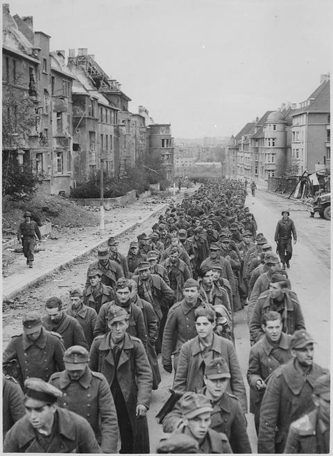 Period photograph: a long column of prisoners in caps filling a street between damaged buildings