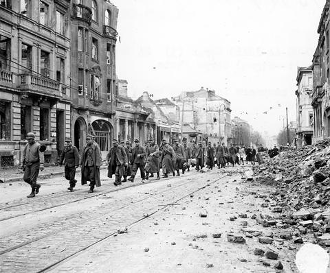 Period photograph: a line of prisoners walking down a wide street with tram tracks and rubble, tall buildings on the left