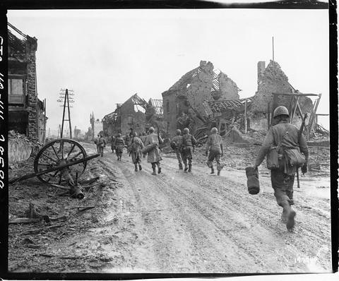 Period photograph: a file of soldiers walking along a muddy village road between ruined houses