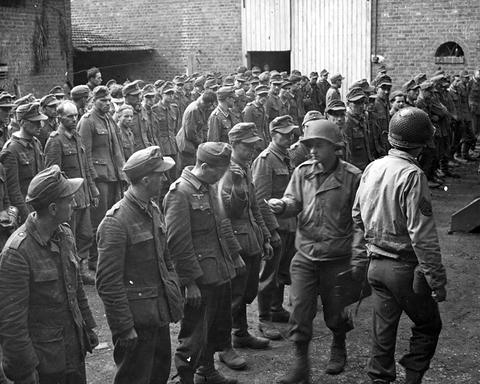 Period photograph: a crowd of prisoners in caps and helmets standing in front of a building, American soldiers beside them