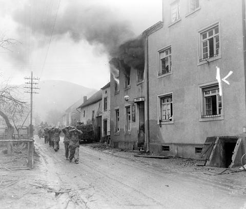 Period photograph: prisoners walking along a village street, smoke pouring from a building on the left