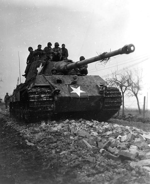 Period photograph: a large tank with a long gun and a white star, men sitting on top, driving past rubble