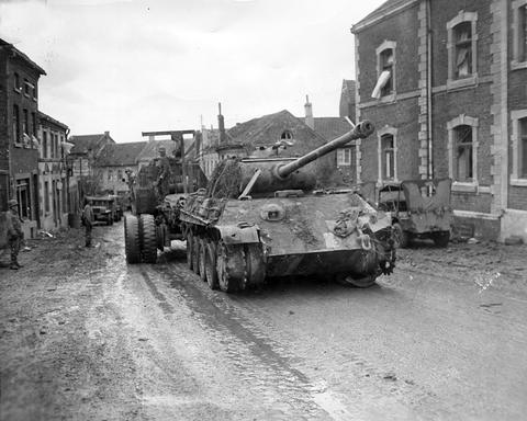 Period photograph: a recovery vehicle towing a tank without tracks along a town street, brick houses on both sides
