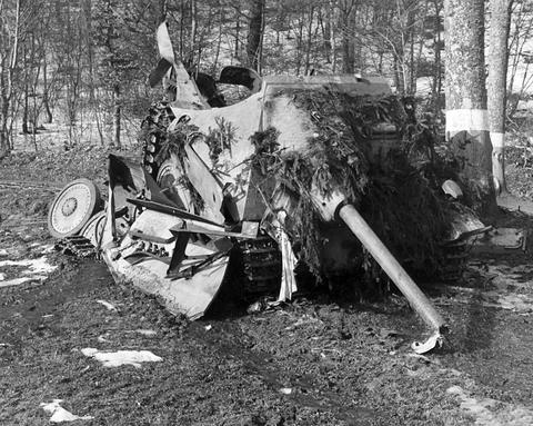 Period photograph: a wrecked tank destroyer tipped on its side by a road, covered with branches, its gun pointing down