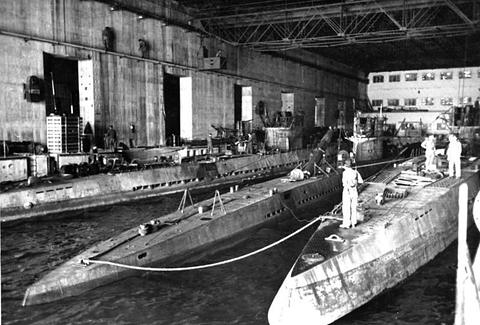 Period photograph: several submarines moored side by side in a long concrete pen, men standing on their decks
