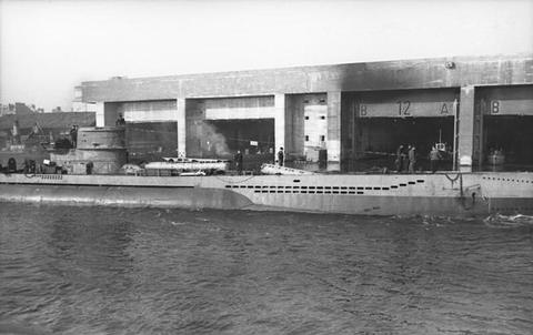 Period photograph: a long, low concrete building with dark openings at the waterline, seen across a basin