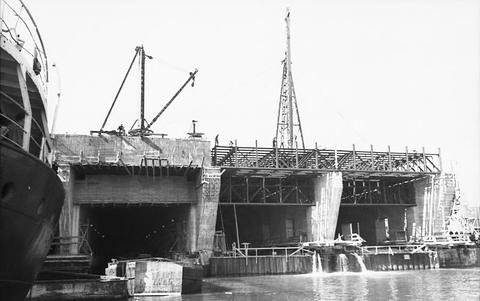 Period photograph: a large building site with cranes, scaffolding and concrete walls beside the water
