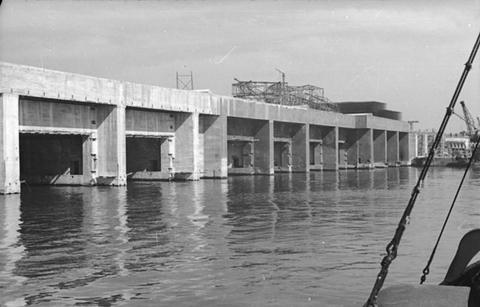 Period photograph: a row of tall concrete bays opening onto the water, scaffolding along the top
