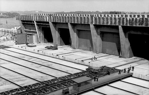 Period photograph: a vast flat concrete roof seen from above, small figures of workers on it, pen bays behind