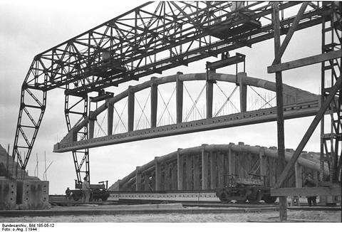 Period photograph: a tall gantry crane lifting a curved steel arch above a vast concrete structure