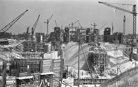 Period photograph: a wide building site with tower cranes and concrete walls, small figures in the distance