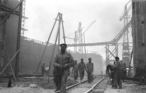 Period photograph: men in striped prisoner clothing walking along rails on a large building site, cranes and concrete walls around them