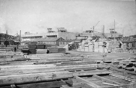 Period photograph: a large building site with stacked timber and boards in front, structures behind