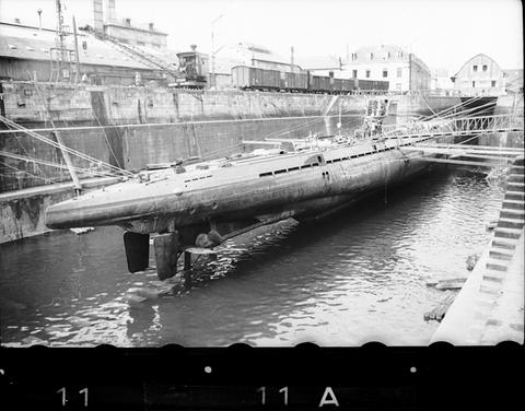 Period photograph: a submarine in a basin beside a quay, buildings behind