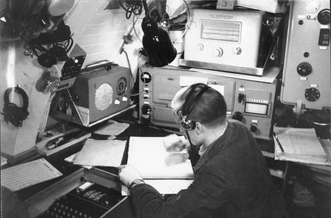 Period photograph: a sailor in a cramped radio room working at a cipher machine with a keyboard