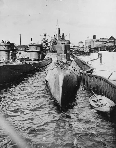 Period photograph: a streamlined submarine moored in a harbour, two other submarines and a cruiser behind