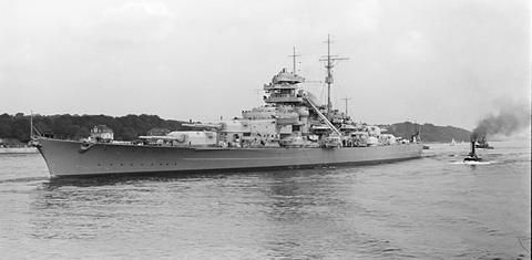 Period photograph: a battleship on a wide river with two tugs at her stern, wooded bank behind