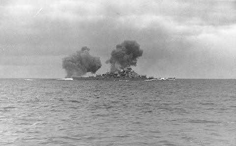 Period photograph: a distant battleship at sea with a flash and smoke from her guns