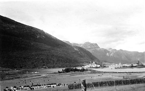 Period photograph: a fjord beneath mountains, warships in the distance and figures along the shore