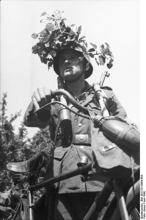 Period photograph: a soldier with a foliage-covered helmet and stick grenades standing with a bicycle