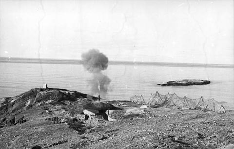 Period photograph: smoke rising from a gun position on a rocky shore above the sea