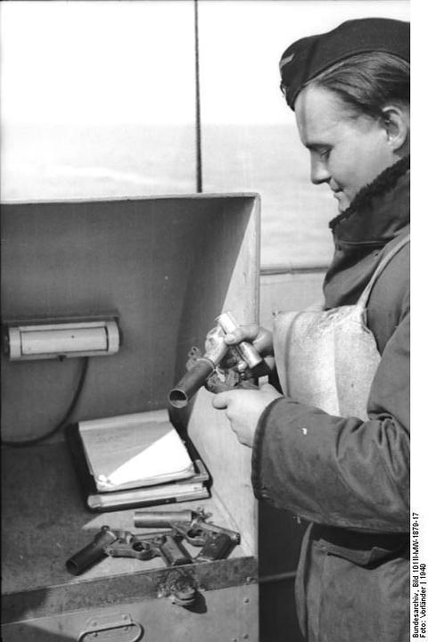 Period photograph: a sailor in a cap and coat holding a flare pistol, sea behind him