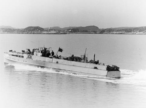 Period photograph: a motor torpedo boat at speed throwing up a bow wave, a low coastline behind