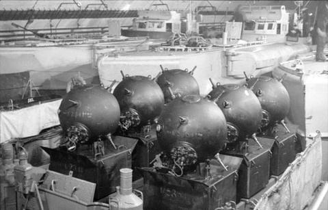 Period photograph: a row of round sea mines on their trolleys on the deck of a boat under a concrete roof