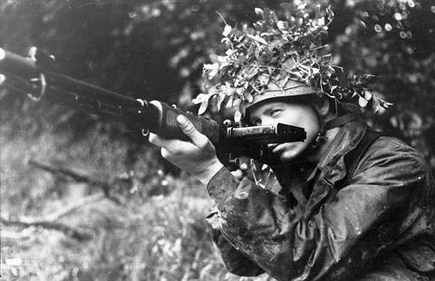 Period photograph: a paratrooper with foliage on his helmet aiming a rifle