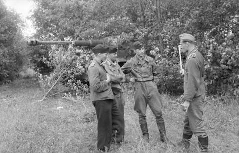 Period photograph: four soldiers standing in a clearing, a tank gun visible among the bushes behind