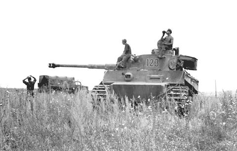 Period photograph: a Tiger tank in tall grass, men on the turret, another soldier in the foreground