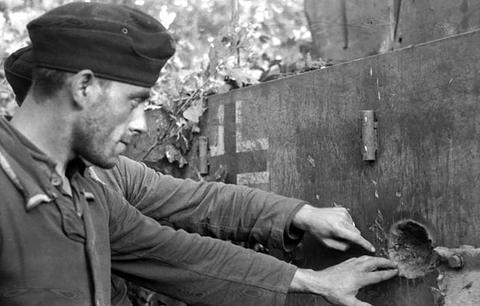 Period photograph: a soldier in a side cap touching a shell scar on thick armour plate