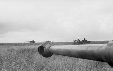 Period photograph: a gun muzzle in the foreground, distant tanks on a flat grass plain