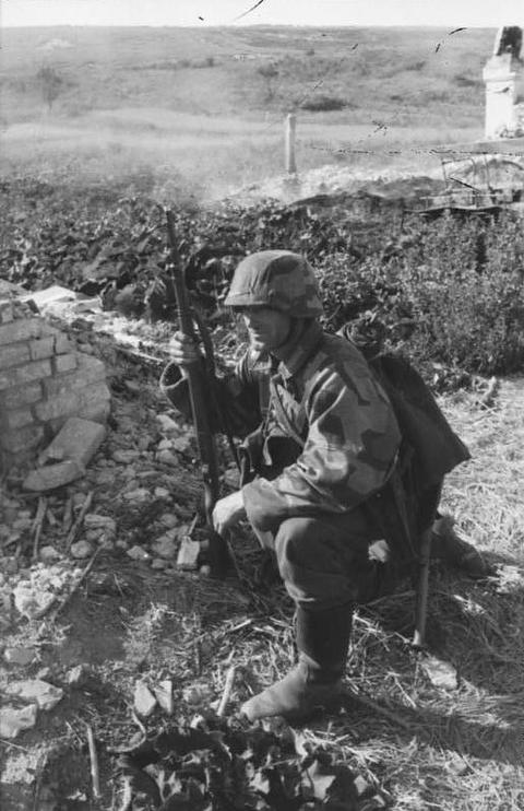 Period photograph: a soldier in a helmet and camouflage jacket kneeling among broken bricks