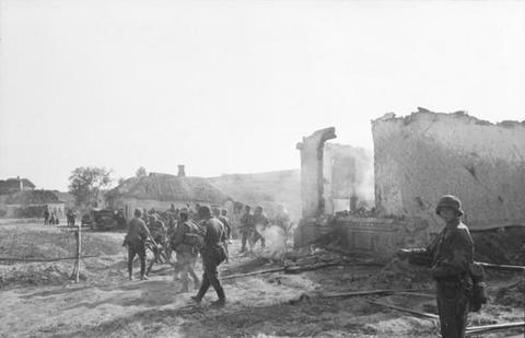 Period photograph: soldiers walking past ruined walls and thatched houses, smoke rising