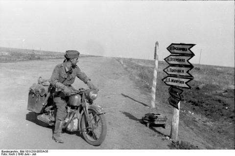 Period photograph: a soldier on a motorcycle beside a post with several direction signs on a flat road