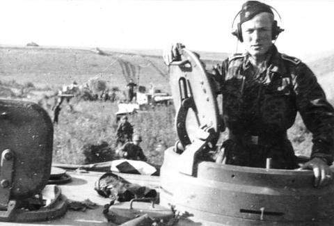 Period photograph: a tank commander in a black cap and headphones standing in a turret hatch