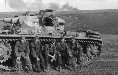 Period photograph: five tank crewmen sitting in front of a tank, smoke rising in the distance