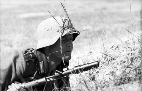 Period photograph: a soldier in a helmet with grass, aiming a rifle with a grenade launcher
