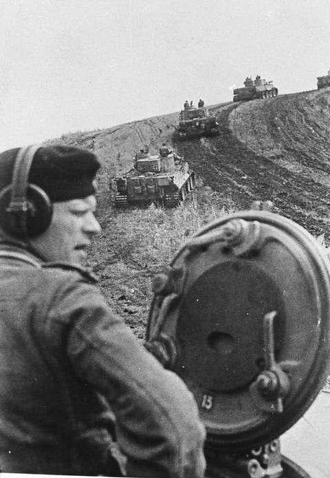 Period photograph: a tank commander in headphones in the foreground, tanks on a slope behind