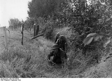 Period photograph: two soldiers crawling through long grass by a fence