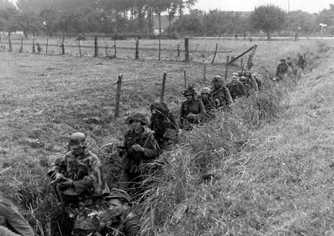 Period photograph: soldiers in camouflage in a ditch beside a fenced meadow