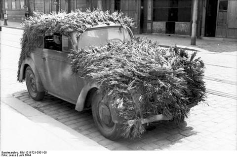 Period photograph: a small car covered in leafy branches on a town street